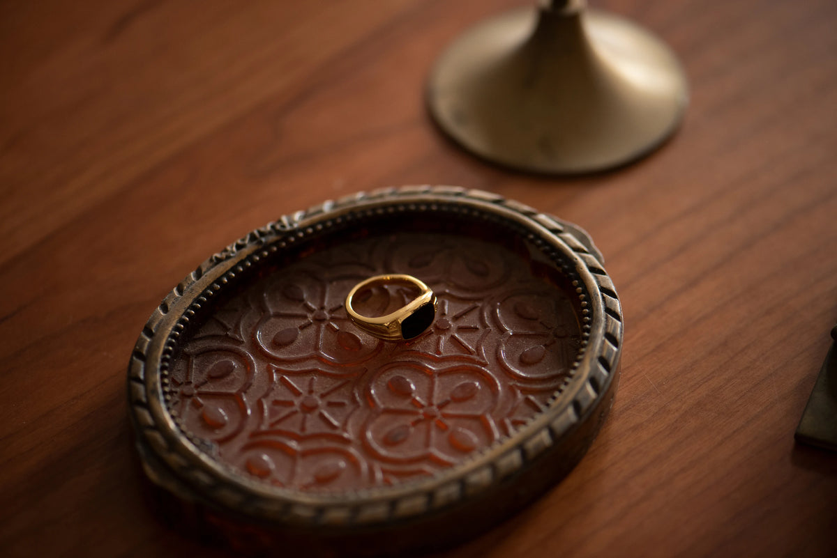 Gold-tone ring with a black rectangular top resting in an ornate oval tray on a wooden table