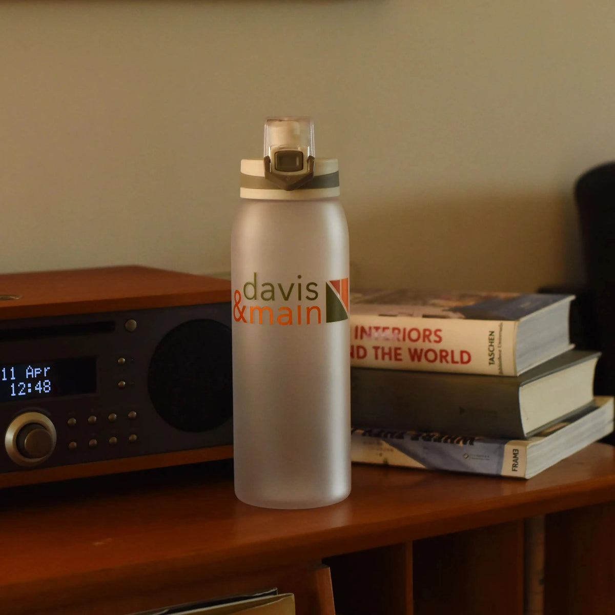 Frosted water bottle with Davis & Main print standing on a wooden shelf beside books and a radio