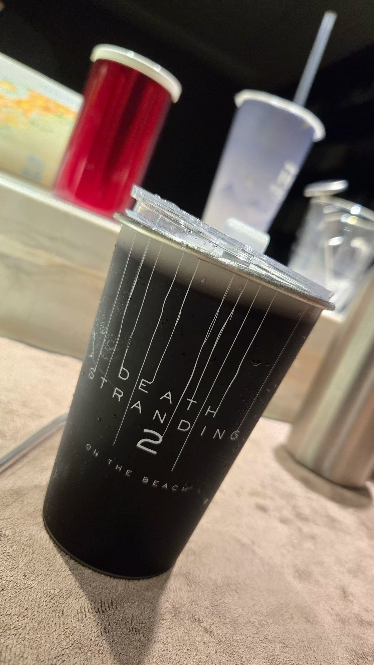 Black tumbler with white vertical line artwork, printed title text and a clear lid on a table